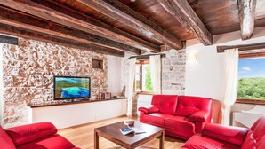 Ein Zimmer mit Steinsäulen, Holzboden und roten Möbeln, inklusive einer Couch und einem Couchtisch. Ein Fernseher ist an der Wand angebracht.