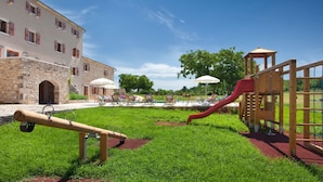 Ein Spielplatz mit Rutsche und Schaukeln, umgeben von einer Rasenfläche und einem Gebäude mit Steinmauern.