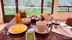 A breakfast table with plates of scrambled eggs, toast, and a beverage, set against a backdrop of a mountainous landscape through large windows. - Boutique Inka Hotel (Ollantaytambo)
