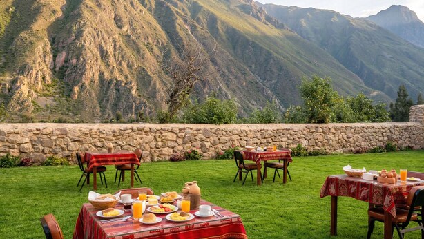 An outdoor dining area with stone walls and tables set for breakfast, overlooking a mountainous landscape. - Boutique Inka Hotel (Ollantaytambo)