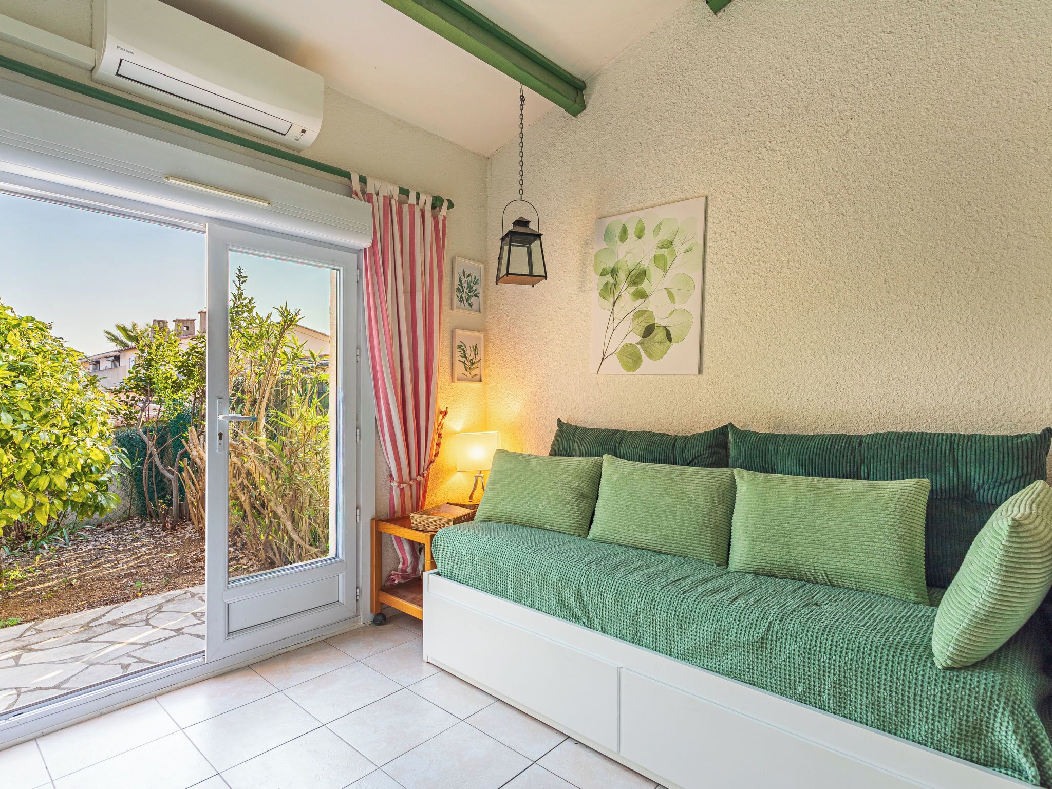 Een kamer met een schuifdeur die uitkomt op een tuin, een groene bank met kussens, een lamp en een wandplant.