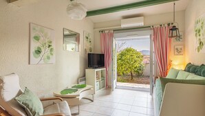 A living room with a TV, a sofa, a chair, a table, and a view of a tree through an open door. - Les Mas de Cogolin by Interhome (Cogolin)