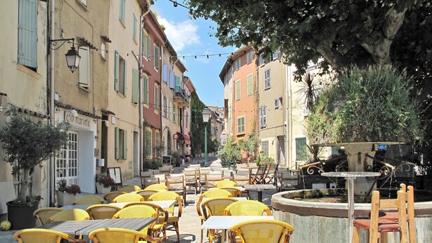 An outdoor café with yellow wicker chairs and tables, surrounded by buildings with balconies and potted plants. - Les Mas de Cogolin by Interhome (Cogolin)