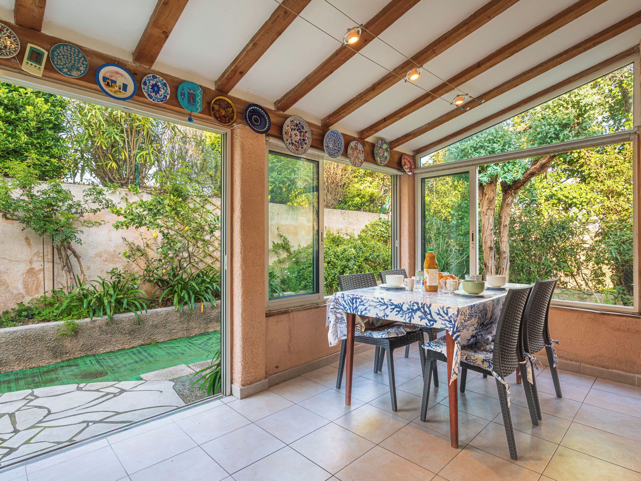 Alpendre coberto com mesa e cadeiras, cercado por grandes janelas com vista para o verde.