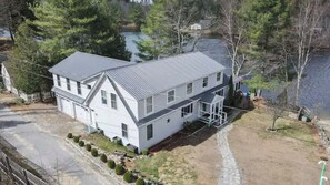 A two-story house with a garage, a covered porch, and a pond in the background. - Impressive, 4 Bedroom Waterfront Home, King Suite (Weare)