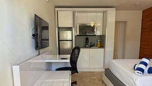 Quarto de hotel compacto com uma cama, escrivaninha, cadeira, micro-ondas e pequena cozinha.