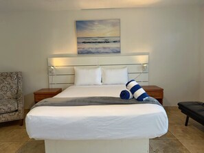 A neatly made bed with a white comforter and pillows, a bedside table with a lamp, a chair, and a painting of a seascape above the bed. - Cozy Beach Studio • Full Kitchen • Pool Access #3 (Pompano Beach)