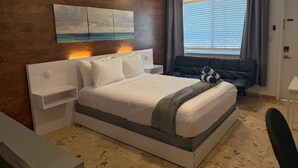 Habitación de hotel con cama, mesitas de noche, escritorio, silla, ventilador de techo, ventana con persianas y un cuadro en la pared.
