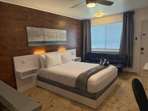 A hotel room with a bed, bedside tables, a desk, a chair, a ceiling fan, a window with blinds, and a painting on the wall. - Warm & Inviting Beach Studio Near Pompano Pier #4 (Pompano Beach)