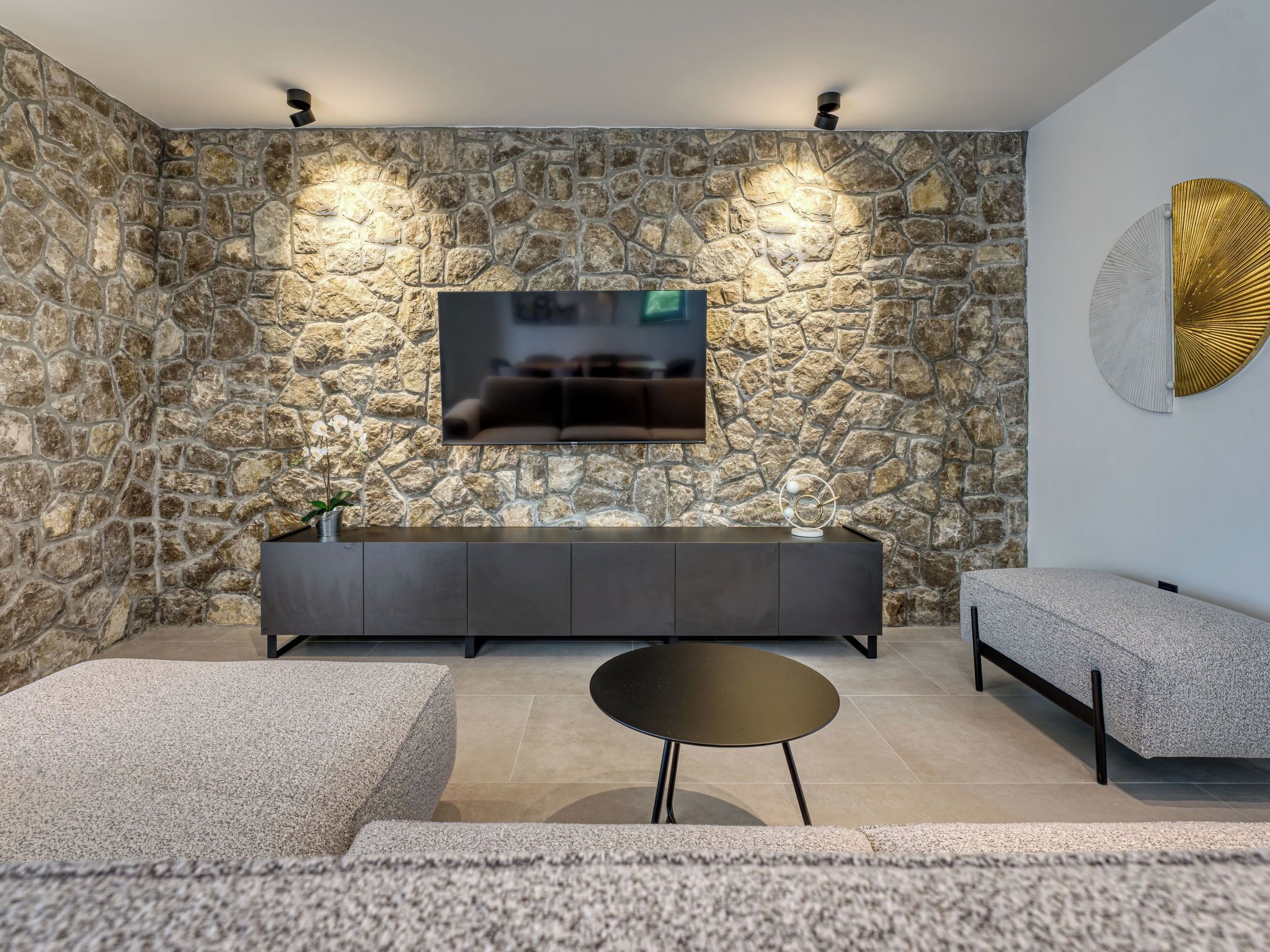 Una sala de estar moderna con una pared de piedra, un televisor de pantalla plana, un sofá oscuro, una mesa redonda y un elemento decorativo en la pared.