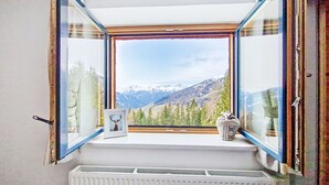 Ein geöffnetes Fenster mit Blick auf Berge, ein Heizkörper darunter und ein gerahmtes Bild auf der Fensterbank.