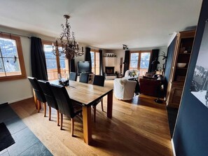 Ein geräumiges Wohnzimmer mit einem Holzessttisch, gemütlicher Sitzgelegenheit, einem Kamin und großen Fenstern mit Blick ins Freie.