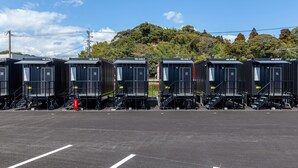 Une rangée de petits bâtiments préfabriqués, chacun doté d’une porte numérotée, est installée sur un parking. Le ciel est dégagé et une colline se profile à l’arrière-plan.