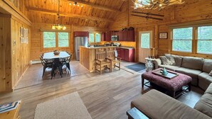 A log cabin interior with a dining area, kitchen, and living room, featuring wooden walls and a ceiling fan. - 7SD Cozy Log Cabin near Bretton Woods with Hot Tub, Firepit, Private Year (Twin Mountain)