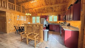 A wooden cabin interior with a dining area, kitchen, and a large window. - 7SD Cozy Log Cabin near Bretton Woods with Hot Tub, Firepit, Private Year (Twin Mountain)