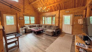 A log cabin interior with a large L-shaped sectional sofa, a chandelier, and multiple windows. - 7SD Cozy Log Cabin near Bretton Woods with Hot Tub, Firepit, Private Year (Twin Mountain)