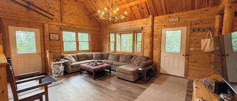 A log cabin interior with a large L-shaped sectional sofa, a chandelier, and multiple windows.