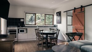 A modern kitchen with white cabinets, a dining table with chairs, a large window with a view of trees, and a sliding barn door. - Rosey Knoll Cottage ~ Cottage with delights of modern life (Daylesford)