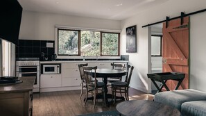 A modern kitchen with white cabinets, a dining table with chairs, a large window with a view of trees, and a sliding barn door. - Rosey Knoll Cottage ~ Cottage with delights of modern life (Daylesford)