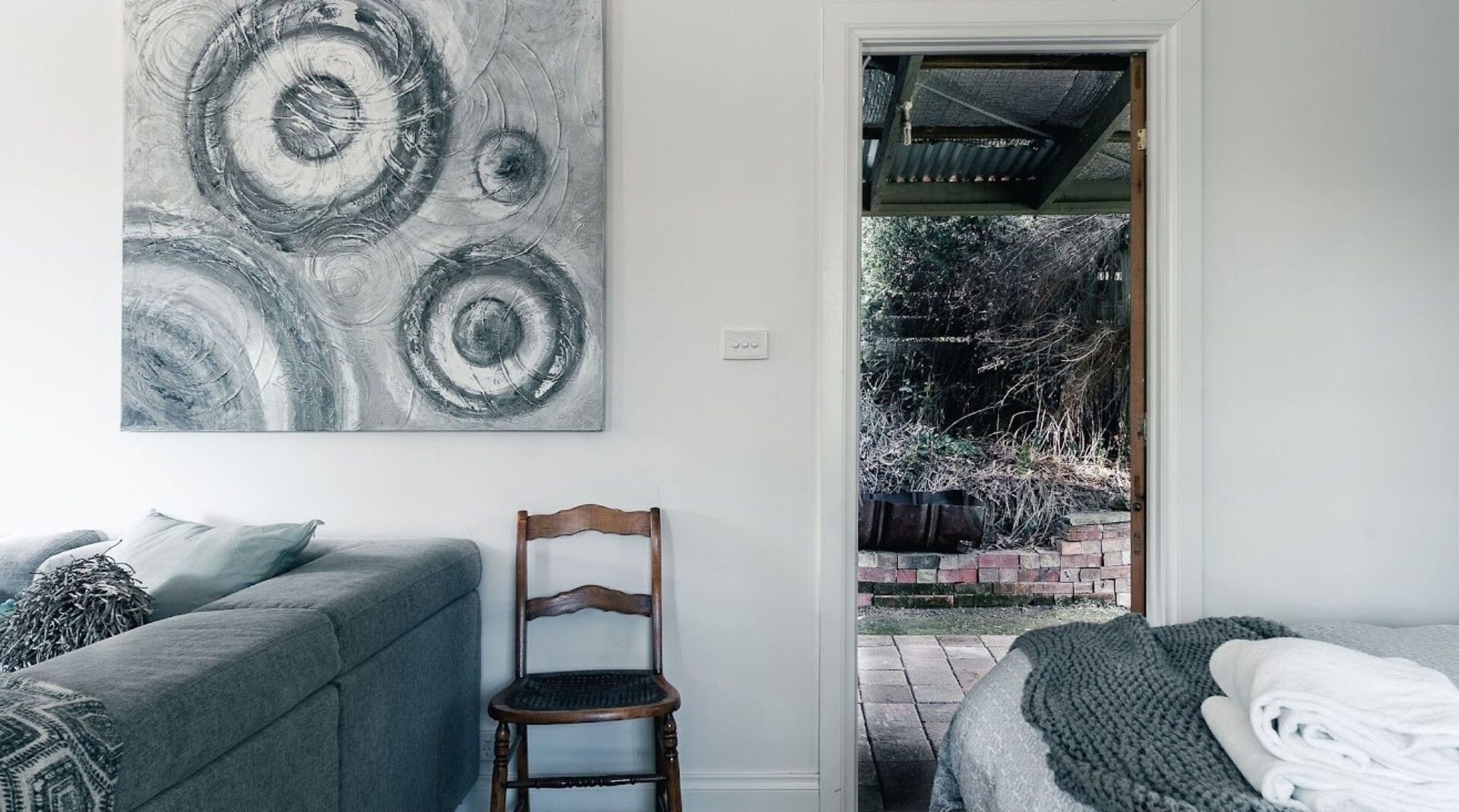 Une pièce avec un canapé gris, une chaise en bois, un grand tableau abstrait et une vue sur un jardin, visible à travers une porte.