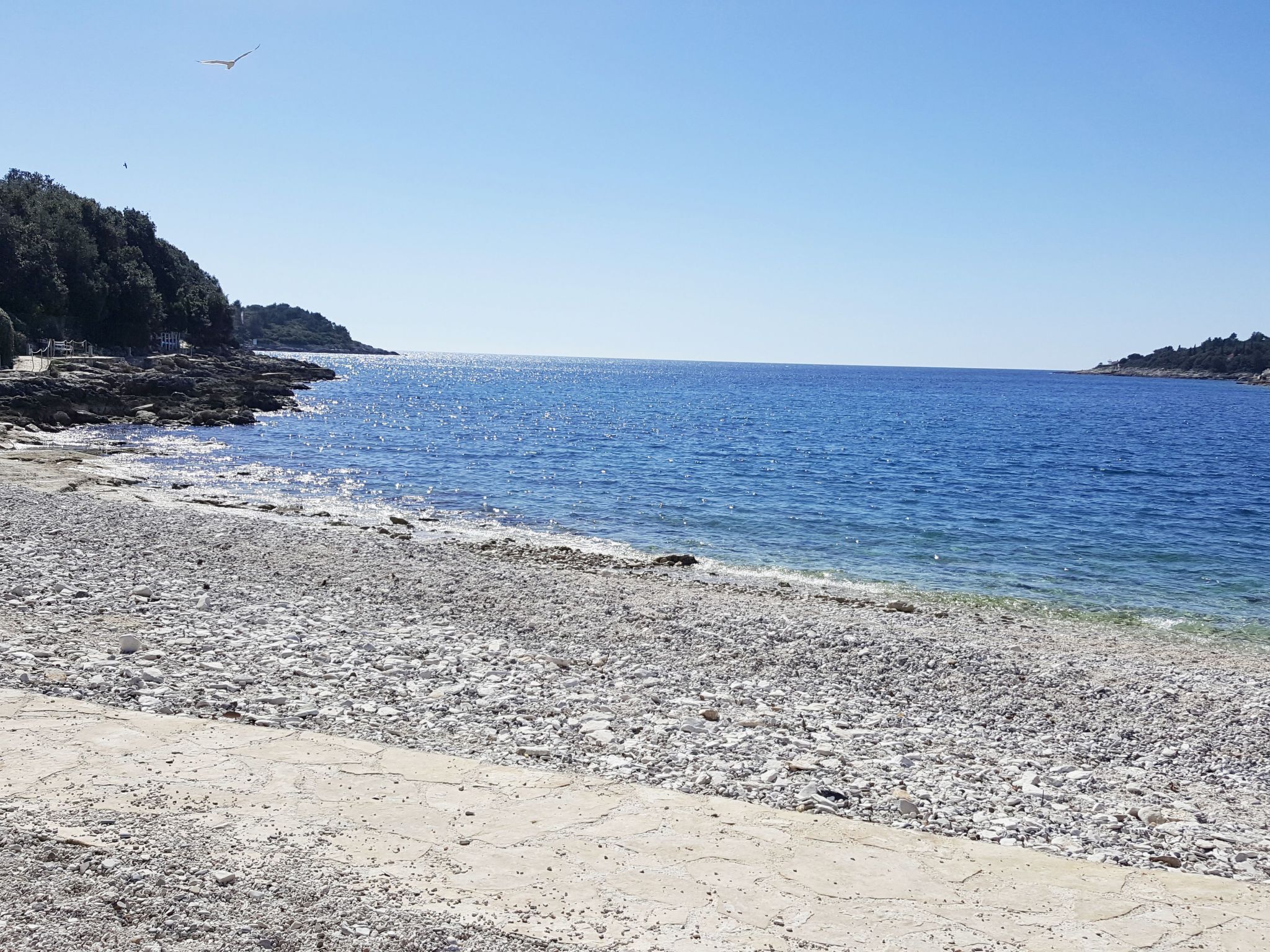 A pebbled beach with clear blue water and rocky cliffs.