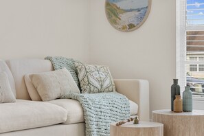 Un salon avec un canapé, des coussins, un plaid en tricot, un tableau accroché au mur et une fenêtre avec des stores.