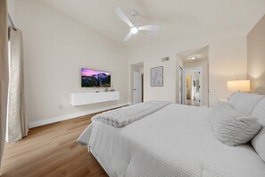 Une chambre à coucher avec un lit, un ventilateur de plafond, un téléviseur fixé au mur et une salle de bain visible à travers une porte ouverte.