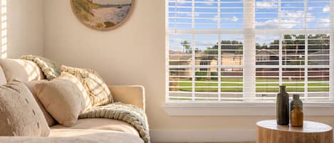 Una habitación con un sofá color beige, un cuadro redondo, una ventana con persianas y una pequeña mesa de madera con dos botellas.