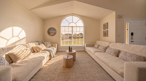 A spacious living room with a high ceiling, a large arched window, and a view of a green landscape. There are two beige sofas and a round wooden coffee table. - 2900 LC - Oak Island Harbor Retreat (Kissimmee)