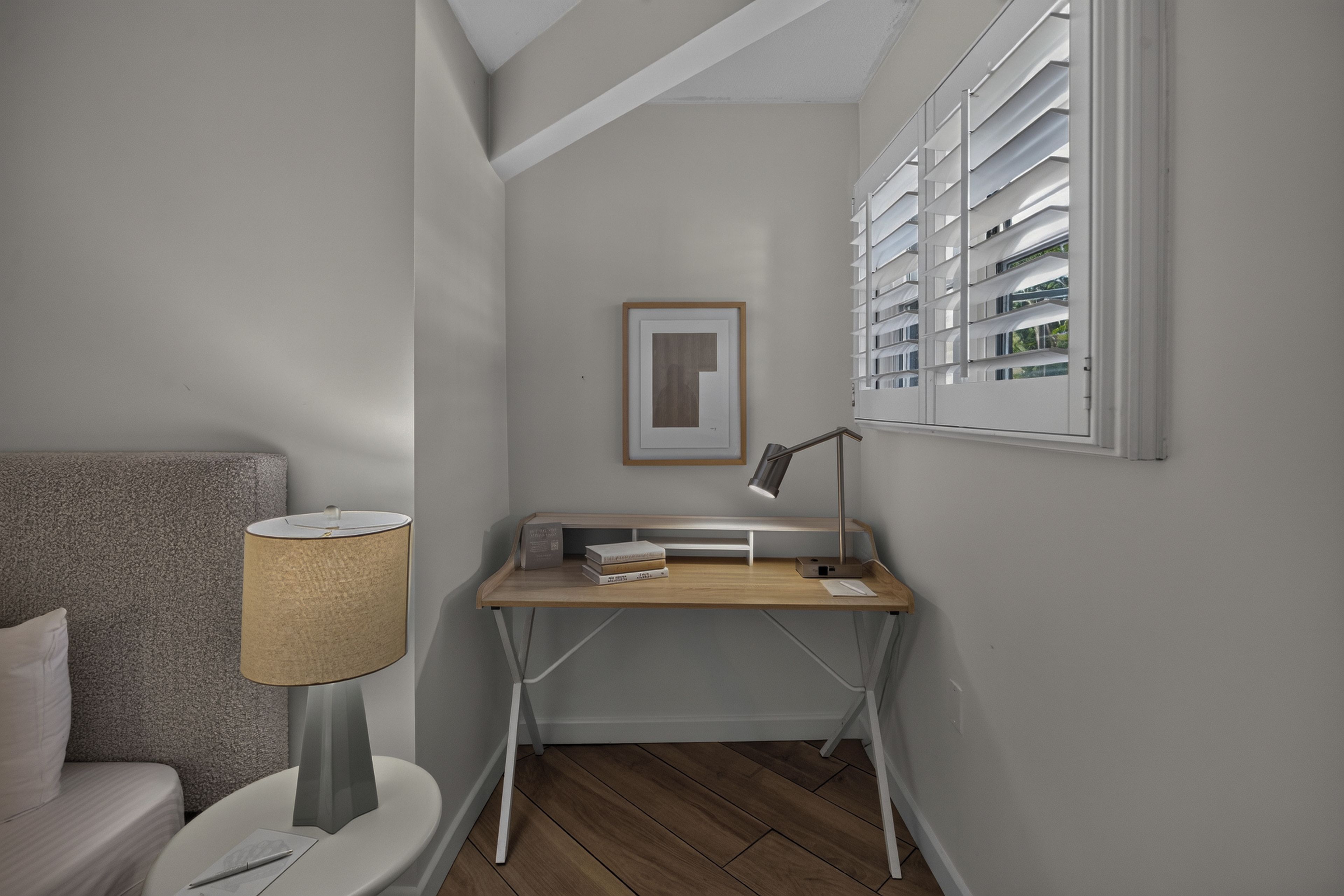 A modern bedroom with a bed, bedside table, lamp, desk, and window with blinds.