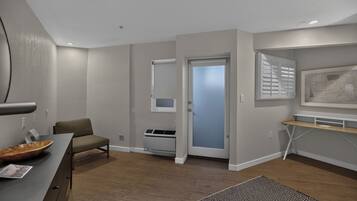 A modern hotel room with a wooden floor, a desk with a chair, a small table, a wall-mounted air conditioner, and a door leading to another room.
