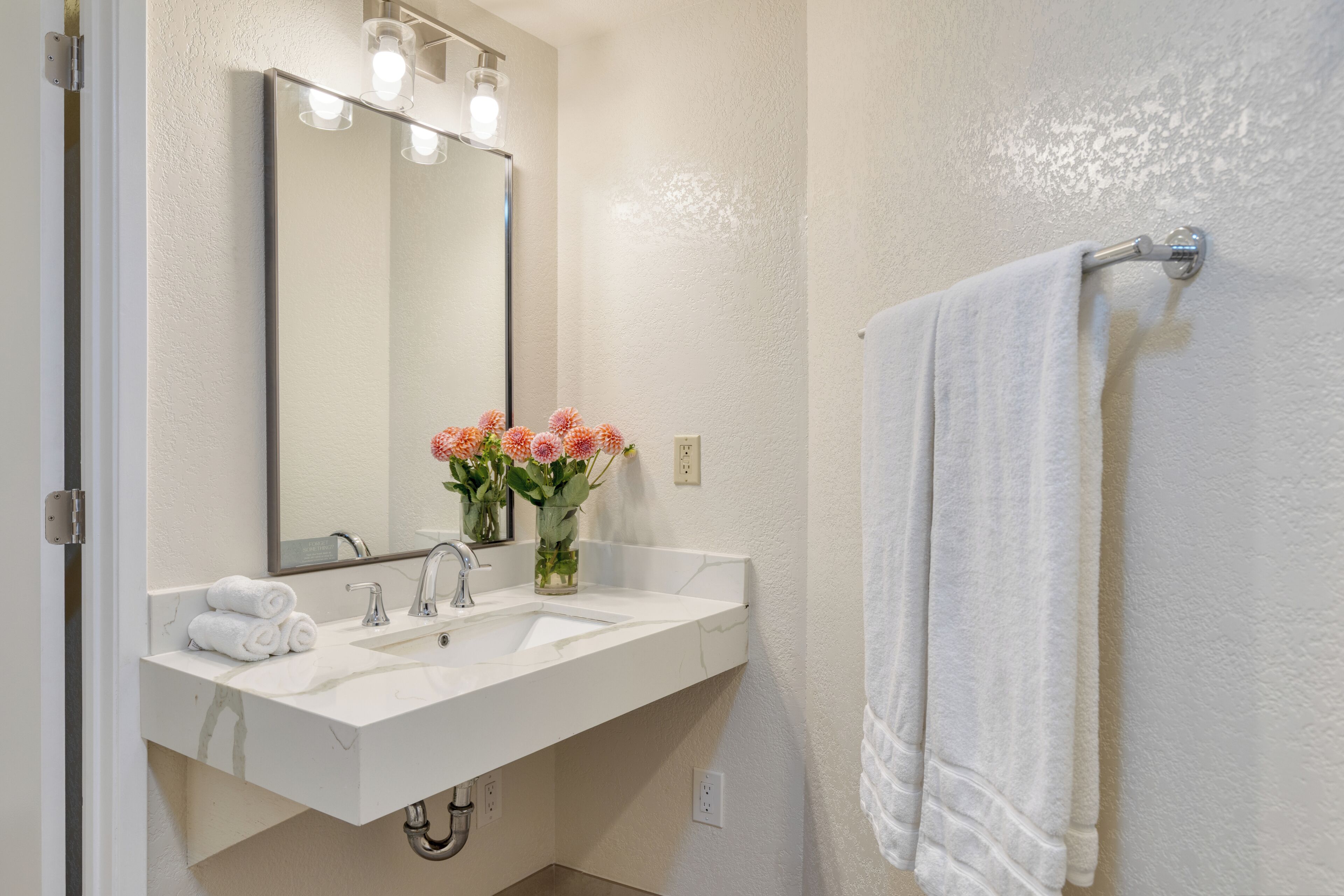 Une salle de bain avec un lavabo blanc, un miroir, un porte-serviettes et un vase rempli de fleurs.