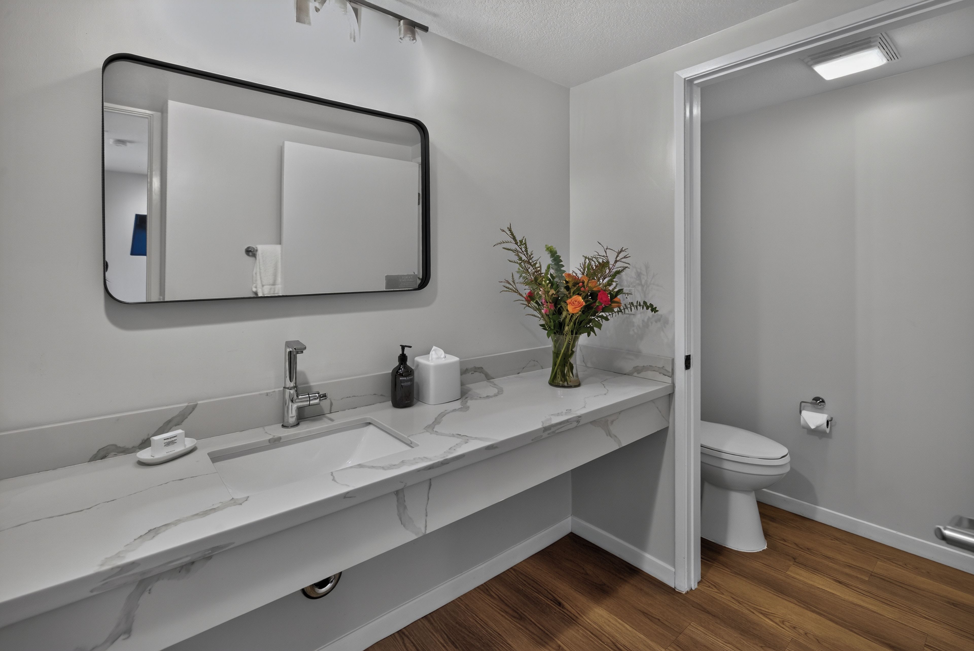 Un baño con tocador blanco, espejo, inodoro y un florero con flores.