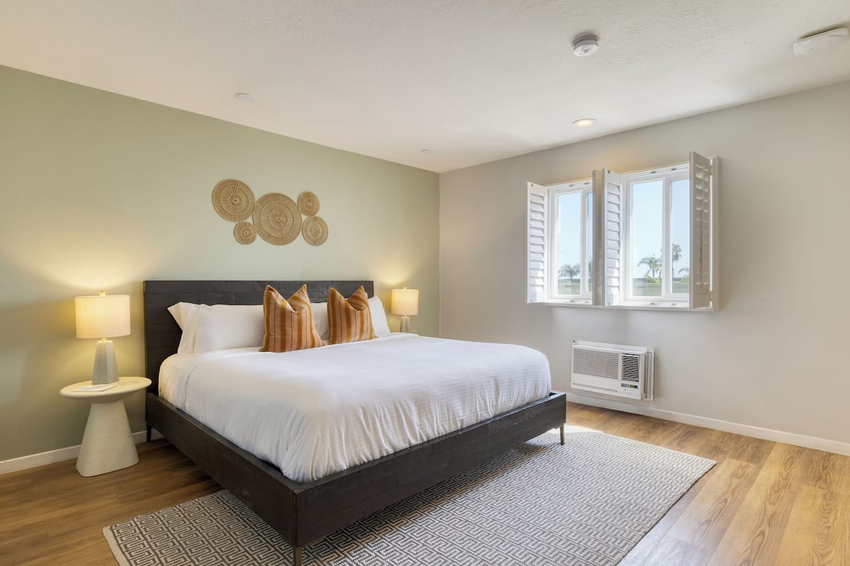 A bedroom with a bed, bedside lamps, a wall-mounted air conditioner, and a window with shutters.