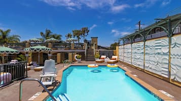 A swimming pool with lounge chairs and umbrellas, surrounded by palm trees and a building with a decorative facade.