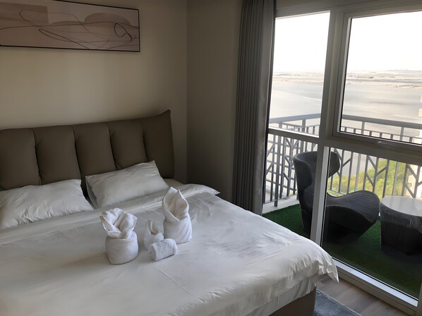 Habitación de hotel con cama, almohadas, ventana que ofrece vistas a un paisaje lejano y balcón con mobiliario exterior.