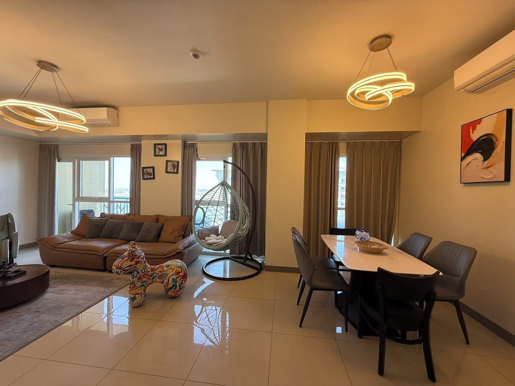 A modern living room with a sofa, a hanging chair, a dining table with chairs, and a large window.