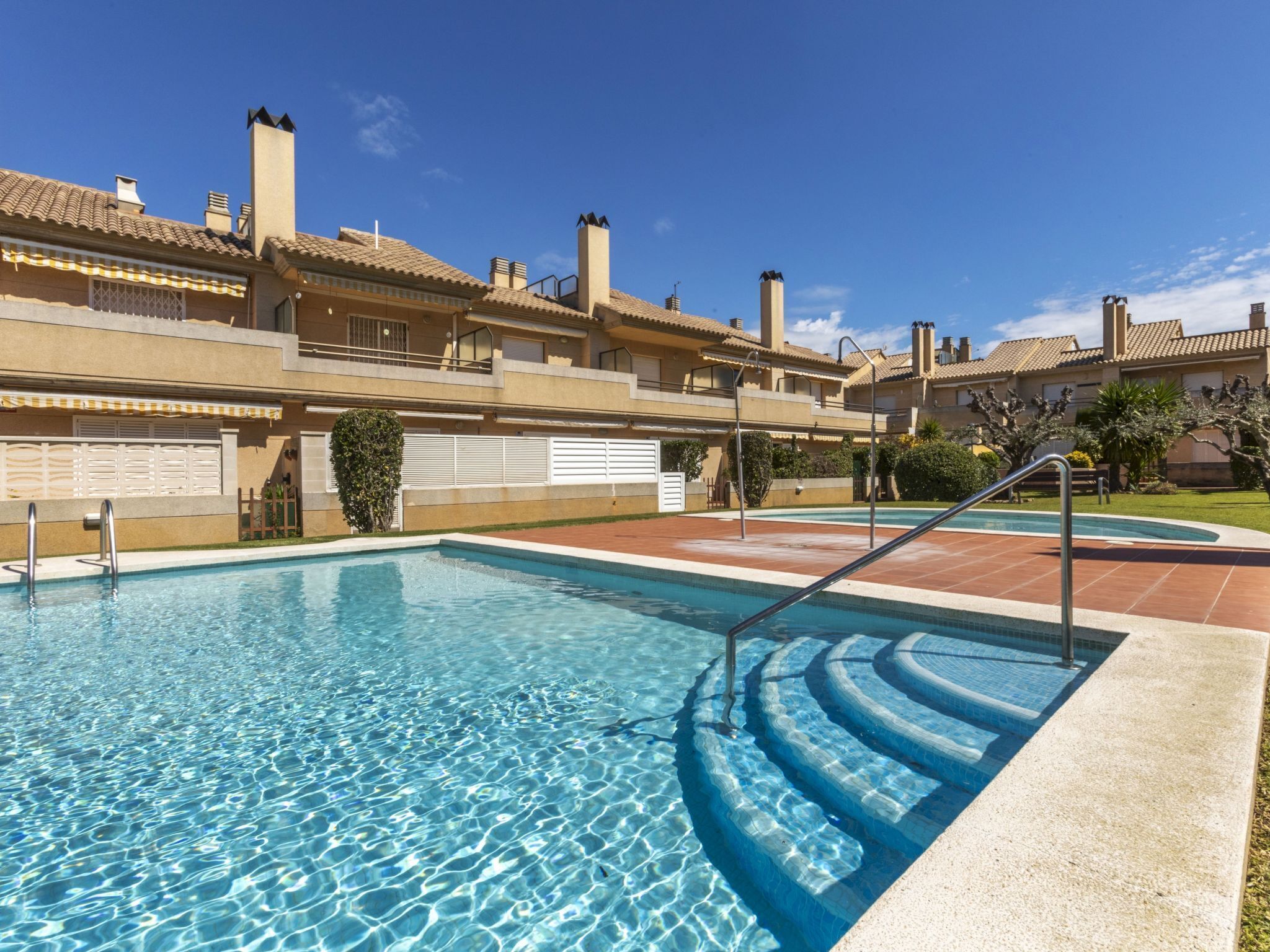 Una piscina con escalones de acceso, rodeada por un edificio con varios balcones y un cielo azul despejado.