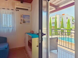 A room with a bed, a desk, an air conditioner, a window with curtains, and a view of a swimming pool and buildings outside. - Villa Roza by Interhome (Umag)