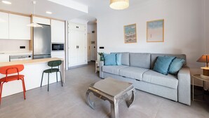 Un salon moderne avec un canapé gris, une petite table et un fauteuil rouge. On y trouve également un coin cuisine avec des meubles blancs et un réfrigérateur.