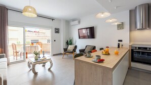 Une cuisine moderne dotée d’un îlot central, un coin repas avec une table et des chaises, et un salon avec un canapé et un téléviseur.