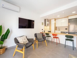 A modern living room with a flat-screen TV, a dining area with colorful chairs, and a kitchen with a stainless steel refrigerator.