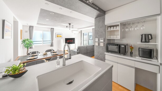 A modern kitchen with a large sink, microwave, and coffee maker. There is a dining area with a table and chairs, and a living room with a sofa and a TV. - ANAX NORD 502 (Sapporo)