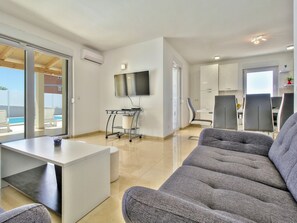 Een moderne woonkamer met een bank, een aan de muur bevestigde televisie, een bijzettafeltje en uitzicht op een zwembad.