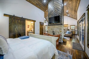 A spacious bedroom with a large bed, a stone fireplace, a wooden ceiling, and a television mounted on the wall. - New Listing Special-Couples Cabin w/Hot Tub, Pool Access, Pickleball & Fire-pit! (Broken Bow)