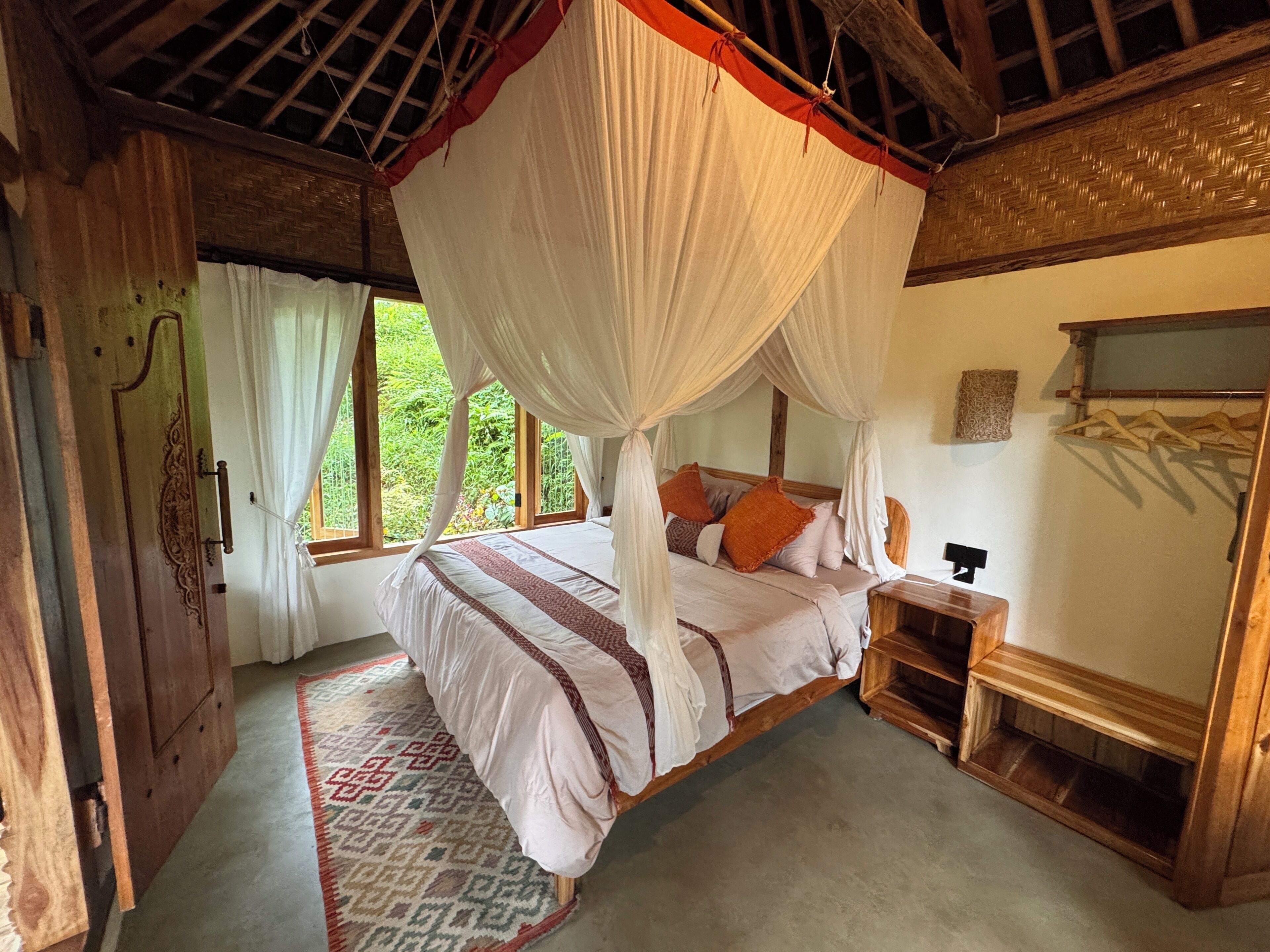 Quarto com cama sob dossel, guarda-roupa de madeira, mesa de cabeceira e vista para o verde lá fora.