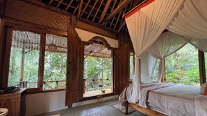 Una stanza con un letto, una panca in legno, una finestra vista su del verde e un baldacchino.