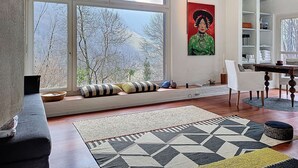 Una habitación con ventanas amplias, un cuadro, un sofá, una mesa de comedor y una estantería.
