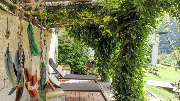 Una terraza de madera con dos sillones de descanso, una jaula colgante y abundante vegetación.
