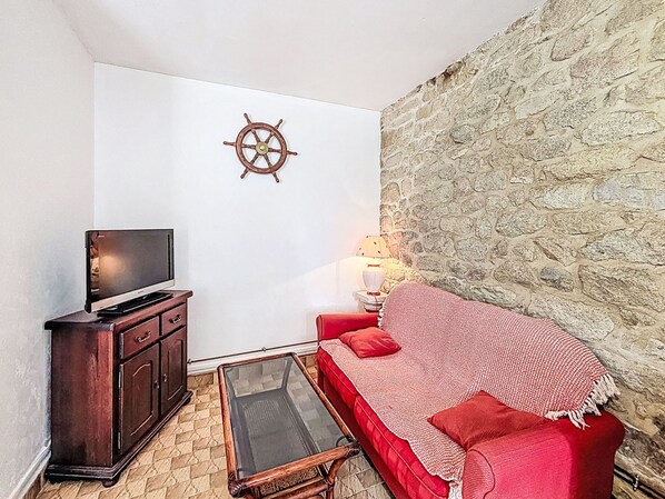 Una sala de estar con una televisión, un aparador de madera, una mesa de centro de vidrio, un sofá rojo, una manta roja y un timón de barco montado en la pared, además de una pared de piedra.